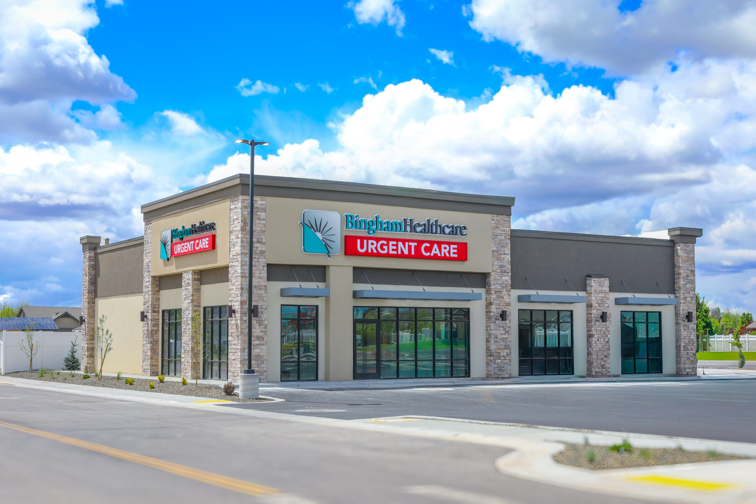 066A3081 1 - Bingham Healthcare Exterior view of a Bingham Healthcare Urgent Care clinic with beige stone facade and large windows against a blue, cloudy sky.